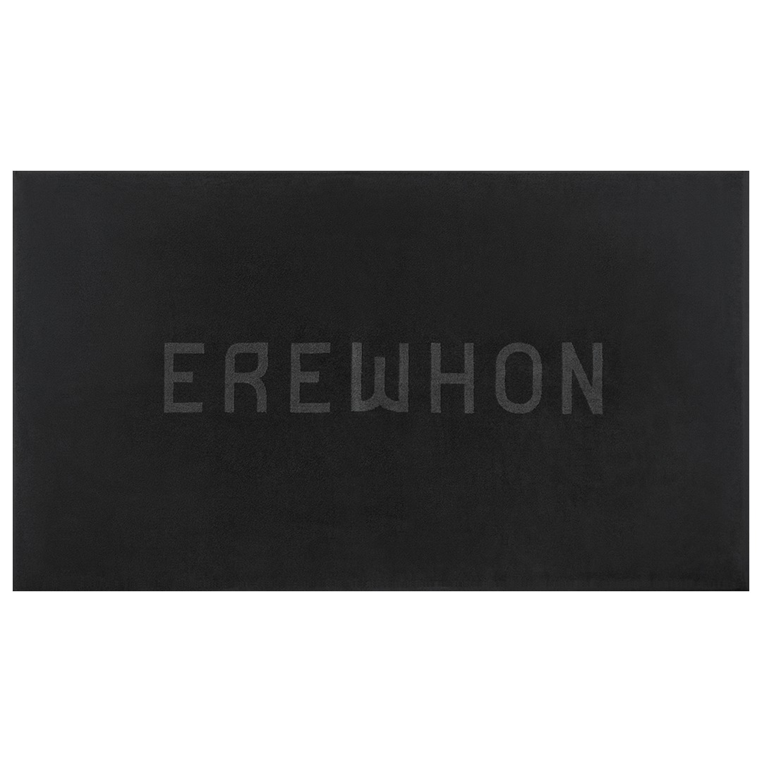 Plush Erewhon Beach Towel in black folded beside its water-resistant travel bag, showcasing the minimalist 3D Jacquard logo in black, apricot, and forest green.